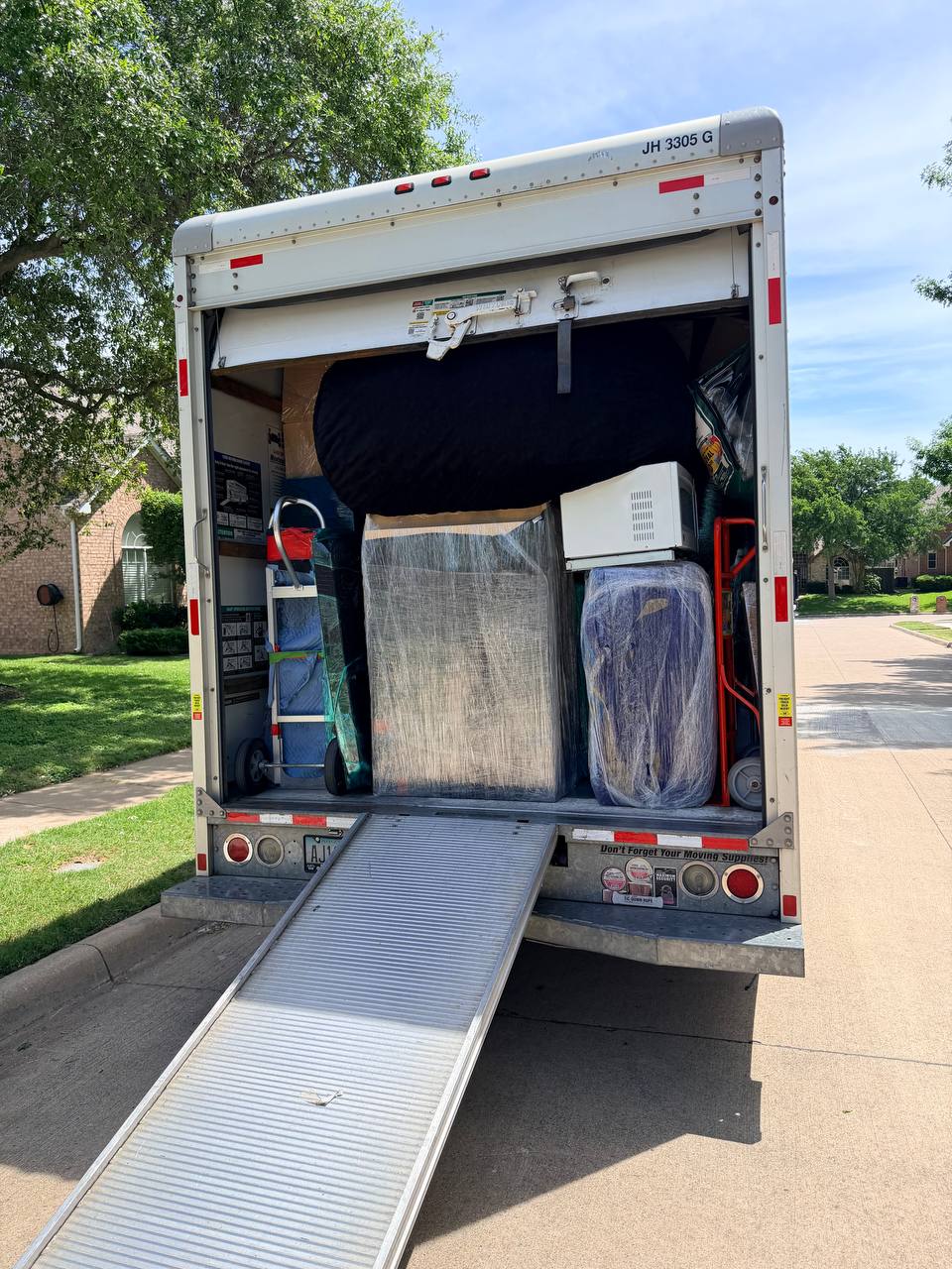Flood Titan box truck loaded with shrink-wrapped customer furniture and contents during a residential pack-out, rear ramp deployed at the curb in a DFW suburban driveway